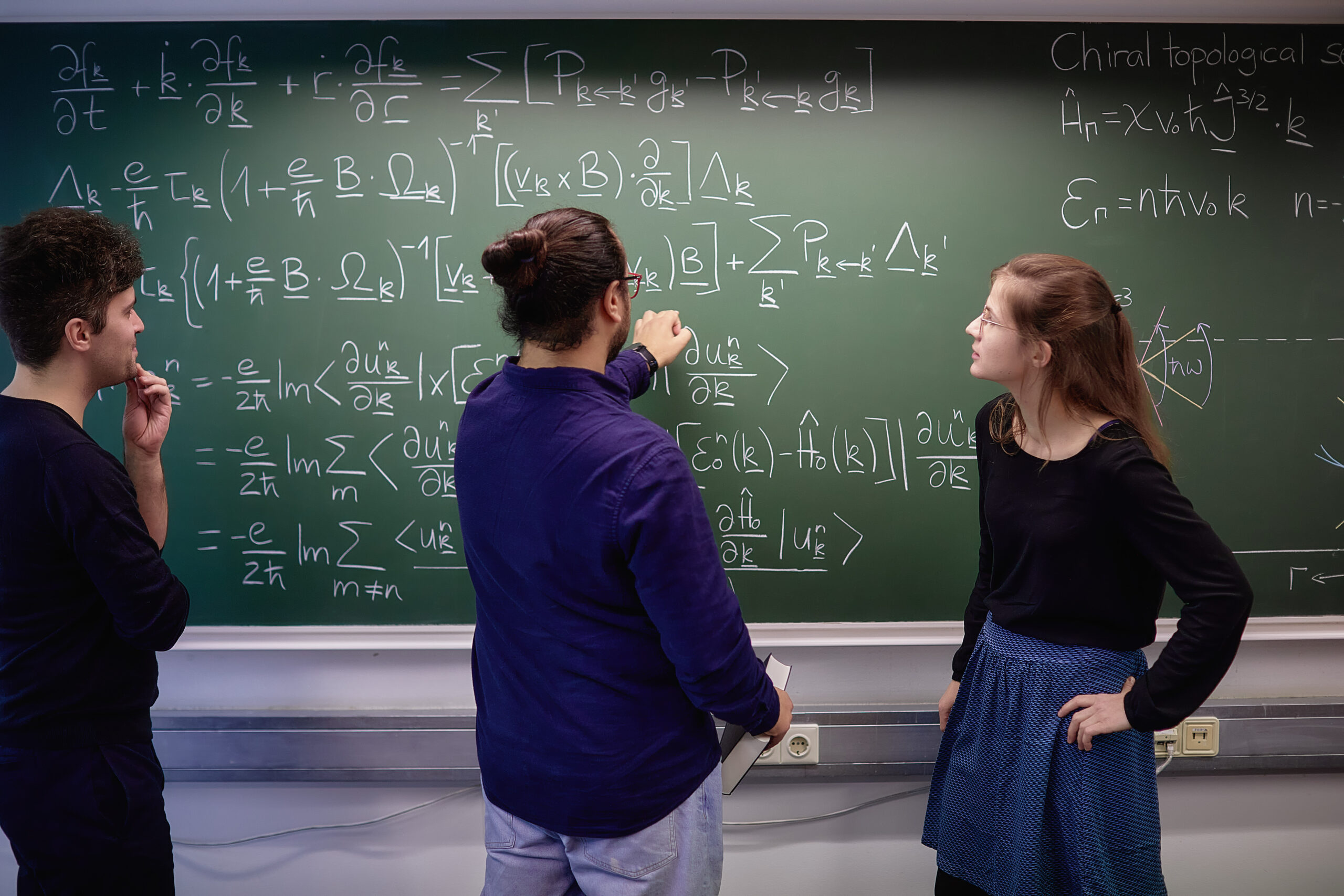 Three researchers discuss equations on chiral topological semimetals at a chalkboard.